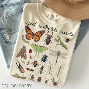 Può includere: Una t-shirt color avorio con la scritta "i just really like insects" in corsivo nero, circondata da una stampa grafica dettagliata di vari insetti, tra cui una farfalla monarca, un cervo volante, una mantide religiosa, una libellula e una falena.