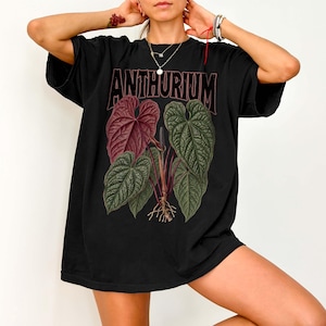 Black unisex oversized t-shirt featuring a detailed Anthurium luxurians botanical illustration with textured dark green and burgundy leaves, roots, and bold “ANTHURIUM” text above, styled as a plant lover graphic tee.