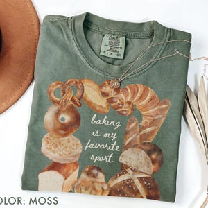 Op de afbeelding: Mosgroen t-shirt met een afbeelding van bakkerijproducten, waaronder pretzels en brood. De tekst "baking is my favorite sport" staat op de voorkant. Een zilveren ketting ligt over het shirt.