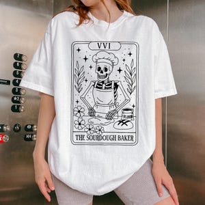May include: White t-shirt featuring a black and white illustration of a skeleton baker wearing a chef's hat and apron. The design includes the text "THE SOURDOUGH BAKER" and the Roman numeral "VVI". The skeleton is depicted baking bread.