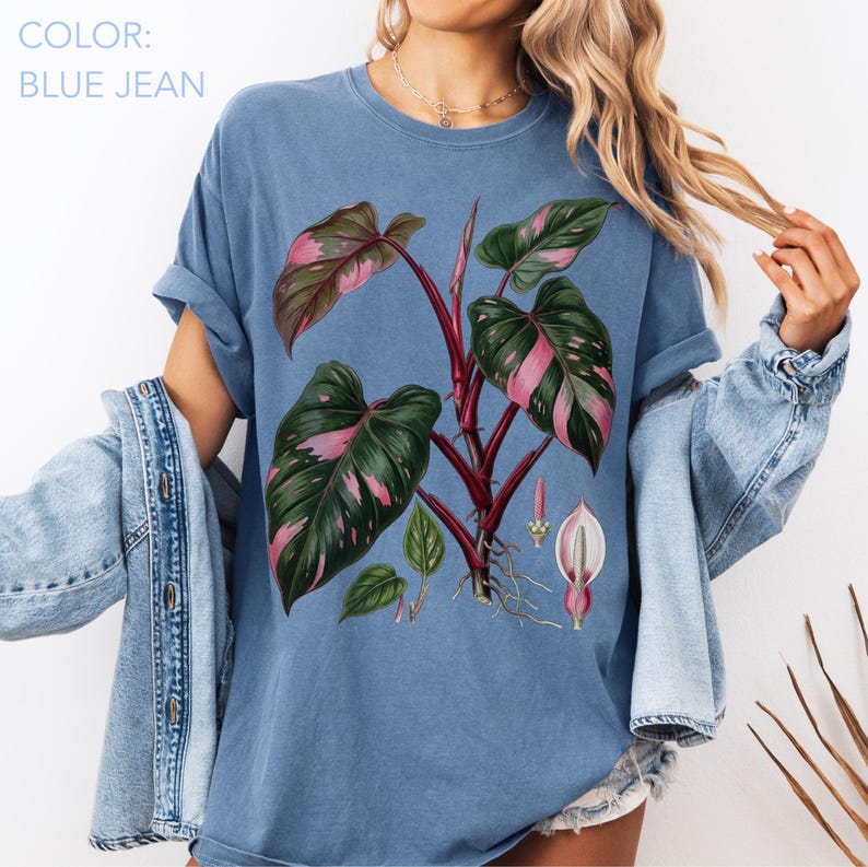 Peut inclure: T-shirt bleu jean avec une illustration botanique d'une plante aux grandes feuilles vertes et aux accents roses. Le t-shirt a des manches retrouss&eacute;es et le texte "COLOR: BLUE JEAN" est visible dans le coin sup&eacute;rieur gauche.