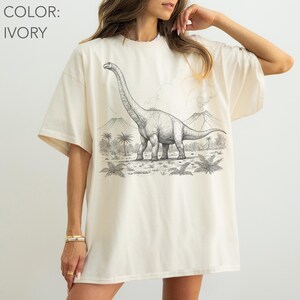 Pu&ograve; includere: T-shirt color avorio con un'illustrazione in bianco e nero di un dinosauro Brachiosaurus in un paesaggio preistorico. Il design include palme e montagne. Il colore della maglietta &egrave; avorio.