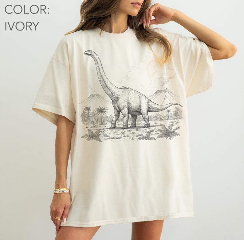 Pu&ograve; includere: T-shirt color avorio con un'illustrazione in bianco e nero di un dinosauro Brachiosaurus in un paesaggio preistorico. Il design include palme e montagne. Il colore della maglietta &egrave; avorio.