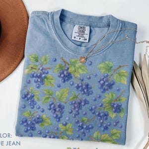 May include: A blue jean colored t-shirt with a grape design. The shirt features a pattern of purple grapes, green leaves, and brown vines. A gold necklace with a pendant is draped across the shirt. The shirt has a Comfort Colors tag.