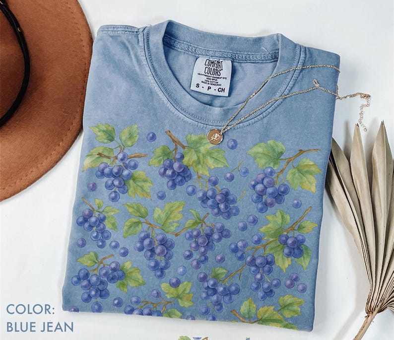 May include: A blue jean colored t-shirt with a grape design. The shirt features a pattern of purple grapes, green leaves, and brown vines. A gold necklace with a pendant is draped across the shirt. The shirt has a Comfort Colors tag.