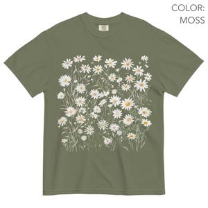 May include: Moss green t-shirt featuring a watercolor design of white daisies with yellow centers and green stems. The shirt has a classic crew neck and short sleeves. The text "COLOR: MOSS" is visible in the upper right corner.