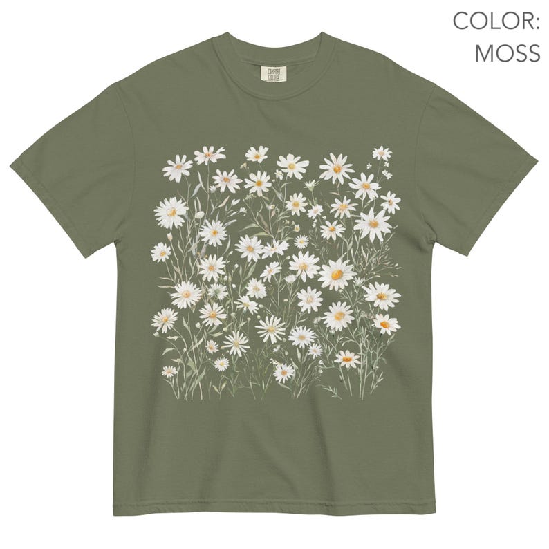 May include: Moss green t-shirt featuring a watercolor design of white daisies with yellow centers and green stems. The shirt has a classic crew neck and short sleeves. The text "COLOR: MOSS" is visible in the upper right corner.