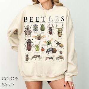 Op de afbeelding: Een zandkleurige sweater met ronde hals en een grafische print met de titel BEETLES COLLECTED CURIOSITIES OF THE NATURAL WORLD. Het ontwerp toont diverse geïllustreerde keversoorten met hun namen in zwarte tekst eronder.