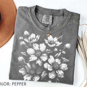 以下が含まれることがあります： 詳細な白い花の模様が施された、ペッパーカラーのTシャツ。Tシャツはクルーネックと半袖です。小さなペンダントが付いたシルバーのネックレスがTシャツにかけられています。Tシャツは白い面に折りたたまれています。