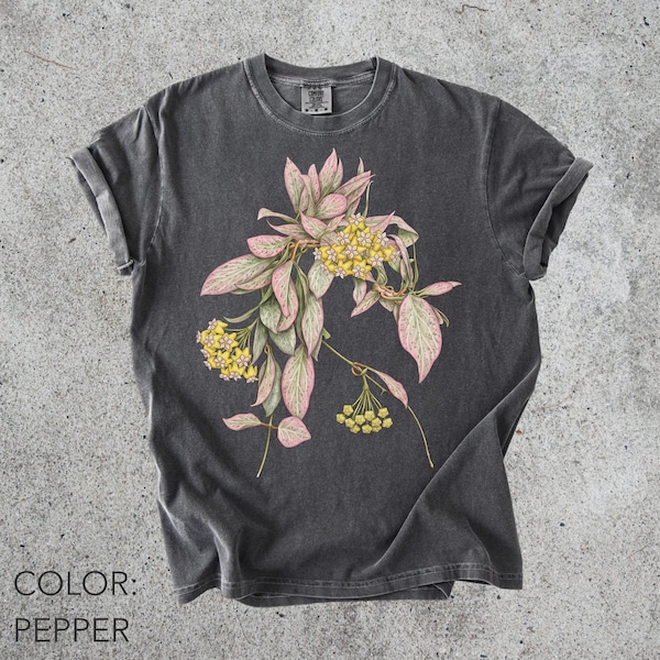 May include: A charcoal gray t-shirt with a vintage-style wash, featuring a central botanical illustration of a plant with variegated pink and green leaves and clusters of small yellow flowers. The shirt has a crew neckline and short sleeves.