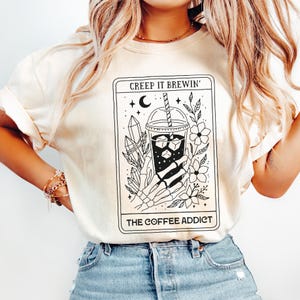 Può includere: T-shirt color crema con grafica in bianco e nero di una bevanda al caffè in stile tarocchi. Il testo recita "Creep It Brewin'" e "The Coffee Addict". Il design include una mano scheletrica, cristalli e fiori.