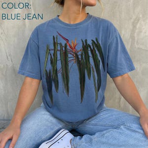 May include: A blue jean colored short-sleeve t-shirt featuring a central graphic print of a botanical illustration. The design displays multiple long, dark green, pointed leaves hanging downward with a single orange and red flower at the top center.