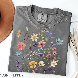 Puede incluir: Una camiseta gris con un dise&ntilde;o floral en tonos rosa, amarillo, azul y morado. La camiseta tiene cuello redondo y mangas cortas. Un collar plateado con un colgante est&aacute; drapeado sobre la camiseta. El texto "COLOR: PEPPER" est&aacute; en la parte inferior.