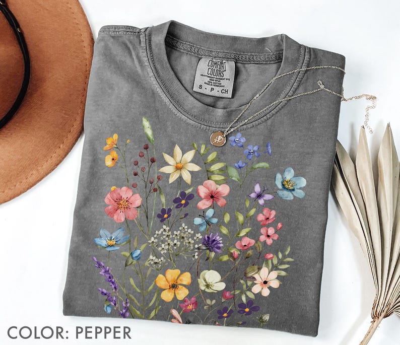 Puede incluir: Una camiseta gris con un dise&ntilde;o floral en tonos rosa, amarillo, azul y morado. La camiseta tiene cuello redondo y mangas cortas. Un collar plateado con un colgante est&aacute; drapeado sobre la camiseta. El texto "COLOR: PEPPER" est&aacute; en la parte inferior.