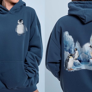 Può includere: Una felpa con cappuccio blu scuro con una piccola grafica di un pulcino di pinguino sul petto. Il retro della felpa mostra un'illustrazione più grande e dettagliata di diversi pinguini in piedi su ghiacciai e neve di colore blu ghiaccio.