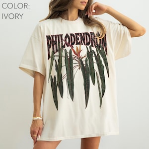 May include: An ivory short-sleeve t-shirt featuring a graphic print of dark green philodendron leaves and the text PHILODENDRON in a bold, distressed black font across the chest. The shirt has a crew neckline and a relaxed, loose-fitting silhouette.
