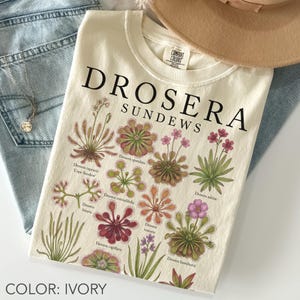以下が含まれることがあります： アイボリー色のTシャツで、様々なドロセラ・サンデュー植物の植物画が描かれており、それぞれの学名が黒い文字で記されています。グラフィックには緑、赤、ピンクの色合いで描かれた様々な種のイラストが含まれており、上部には大きな黒い文字で「DROSERA SUNDEWS」というタイトルが印刷されています。