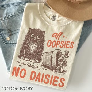 May include: Ivory-colored t-shirt featuring a brown illustration of a fluffy cat, a tipped-over flower pot, and the text "all OOPSIES NO DAISIES" in orange. The shirt is a Comfort Colors brand.