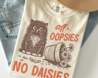 Puede incluir: Camiseta color marfil con una ilustración marrón de un gato esponjoso, una maceta volcada y el texto naranja "all OOPSIES NO DAISIES". Marca Comfort Colors.