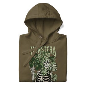 May include: Khaki green hoodie featuring a skeleton design with monstera leaves. The word "MONSTERA" is printed above the skeleton. The hoodie has a drawstring hood and a front pocket. The design is printed on the front.