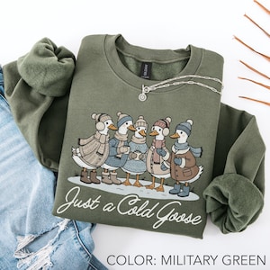 Puede incluir: Sudadera verde militar con un gráfico de cinco gansos vestidos con ropa de invierno. Los gansos llevan sombreros, bufandas y abrigos, con la frase "Just a Cold Goose" en cursiva. Un collar plateado está sobre la sudadera.