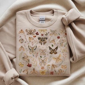 May include: A beige crewneck sweatshirt featuring a detailed graphic print of various moths and butterflies in shades of brown, tan, and cream, interspersed with small botanical illustrations of dried flowers, leaves, and stems.