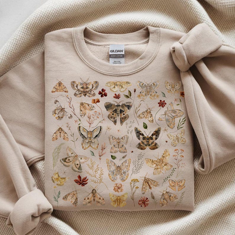 May include: A beige crewneck sweatshirt featuring a detailed graphic print of various moths and butterflies in shades of brown, tan, and cream, interspersed with small botanical illustrations of dried flowers, leaves, and stems.