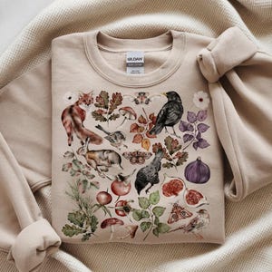 Può includere: Felpa beige con un'illustrazione ad acquerello di animali della foresta, piante e insetti. Il design include una volpe, un coniglio, uccelli, funghi e falene. La felpa ha un girocollo e maniche lunghe.