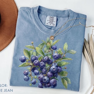 May include: Blue jean coloured t-shirt featuring a watercolour illustration of blueberries and green leaves. The shirt has a crew neck and short sleeves. A gold necklace with a small pendant is draped across the shirt.