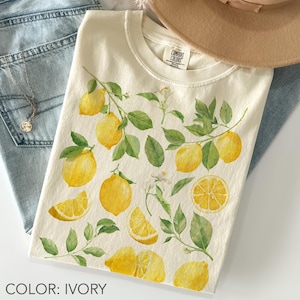 May include: Ivory-colored t-shirt featuring a watercolor design of lemons, leaves, and blossoms. The design includes whole lemons, lemon slices, and sprigs of leaves. The shirt is paired with blue jeans and a necklace.