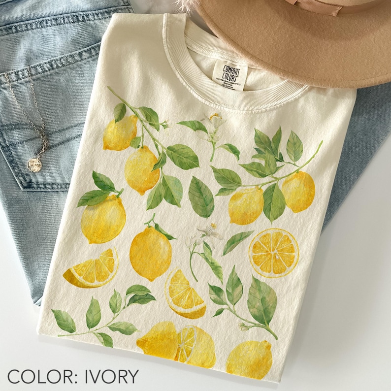 May include: Ivory-colored t-shirt featuring a watercolor design of lemons, leaves, and blossoms. The design includes whole lemons, lemon slices, and sprigs of leaves. The shirt is paired with blue jeans and a necklace.