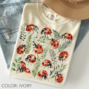 Peut inclure: T-shirt ivoire avec un motif aquarelle de coccinelles rouges à pois noirs et de feuillage vert. Le t-shirt a un col rond et des manches courtes. Le texte "COLOR: IVORY" est au bas de l'image.