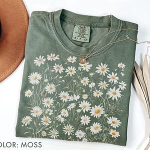 May include: Moss green t-shirt with a daisy floral design. The shirt has a crew neck and short sleeves. A gold necklace with a pendant is draped over the shirt. The shirt is labeled "Comfort Colors".