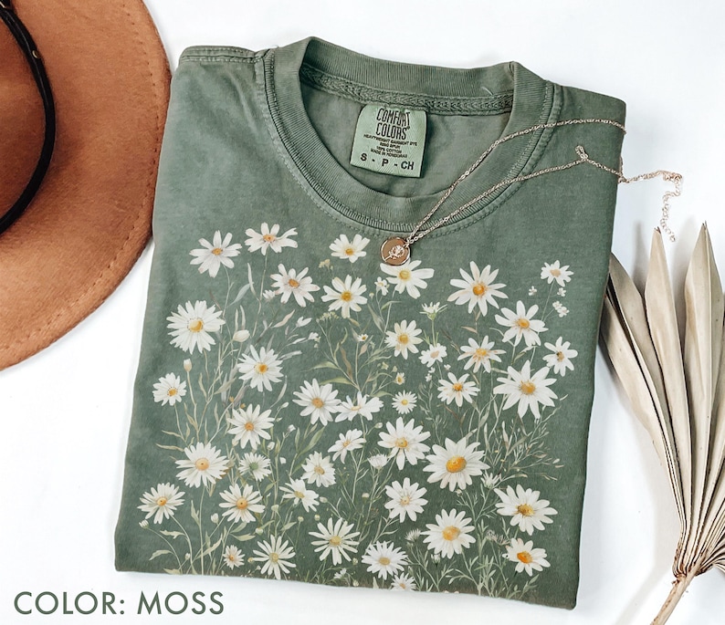 May include: Moss green t-shirt with a daisy floral design. The shirt has a crew neck and short sleeves. A gold necklace with a pendant is draped over the shirt. The shirt is labeled "Comfort Colors".