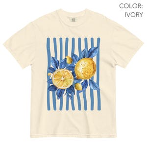 May include: Ivory-colored t-shirt featuring a watercolor design of lemons and blue leaves. The design is set against a backdrop of vertical blue stripes. The shirt has a classic crew neck and short sleeves.