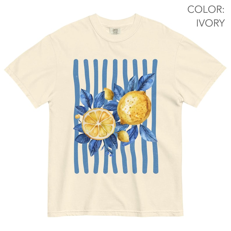 May include: Ivory-colored t-shirt featuring a watercolor design of lemons and blue leaves. The design is set against a backdrop of vertical blue stripes. The shirt has a classic crew neck and short sleeves.