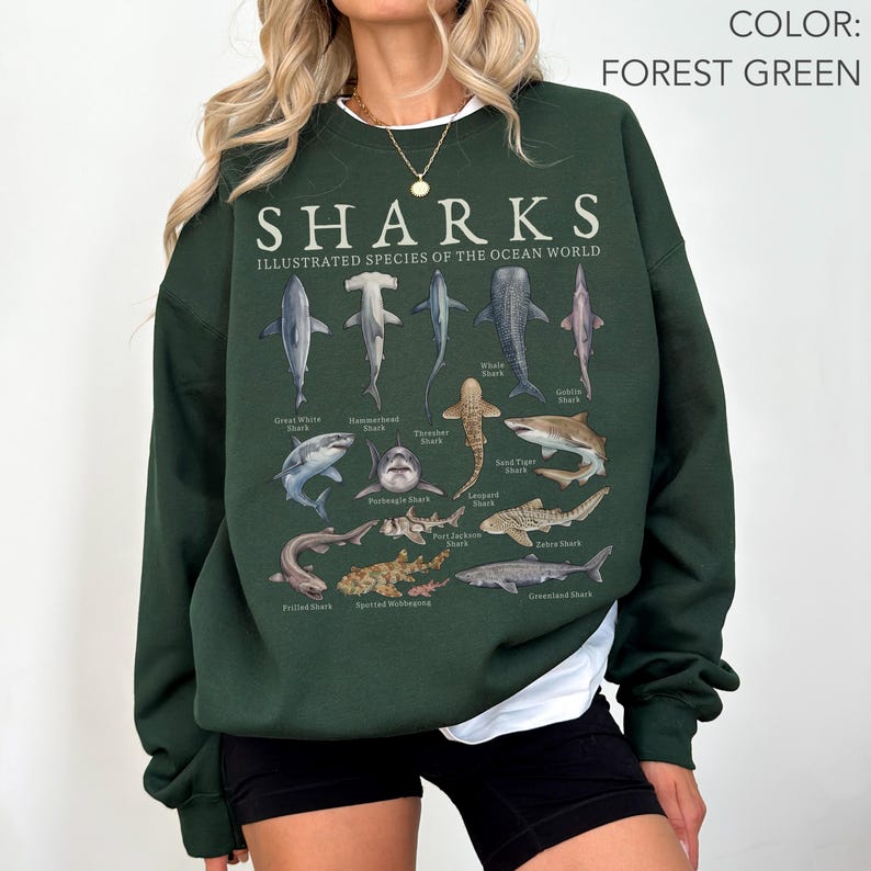 Woman wearing a forest green Gildan 18000 sweatshirt featuring a labeled vintage shark species chart with great white, hammerhead, whale shark, goblin shark, thresher shark, and more.