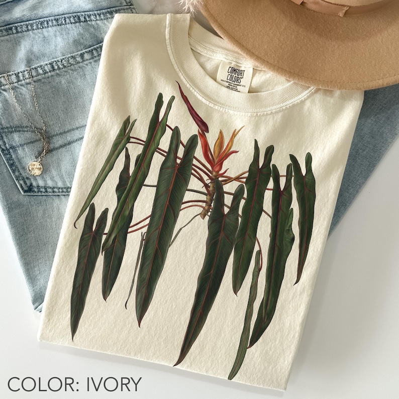 Folded ivory Comfort Colors T-shirt featuring a Philodendron spiritus-sancti with long dark green leaves, red stems and a vibrant tropical inflorescence.