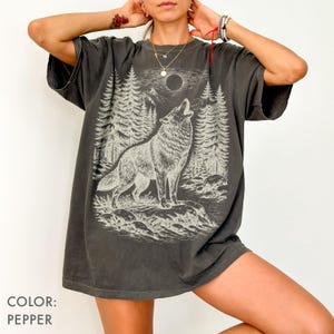 Puede incluir: Una camiseta color pimienta con un estampado gr&aacute;fico de un lobo aullando a la luna llena en un entorno forestal. El dise&ntilde;o incluye ilustraciones detalladas de pinos, terreno rocoso y una luna circular, representadas en tonos claros y apagados.