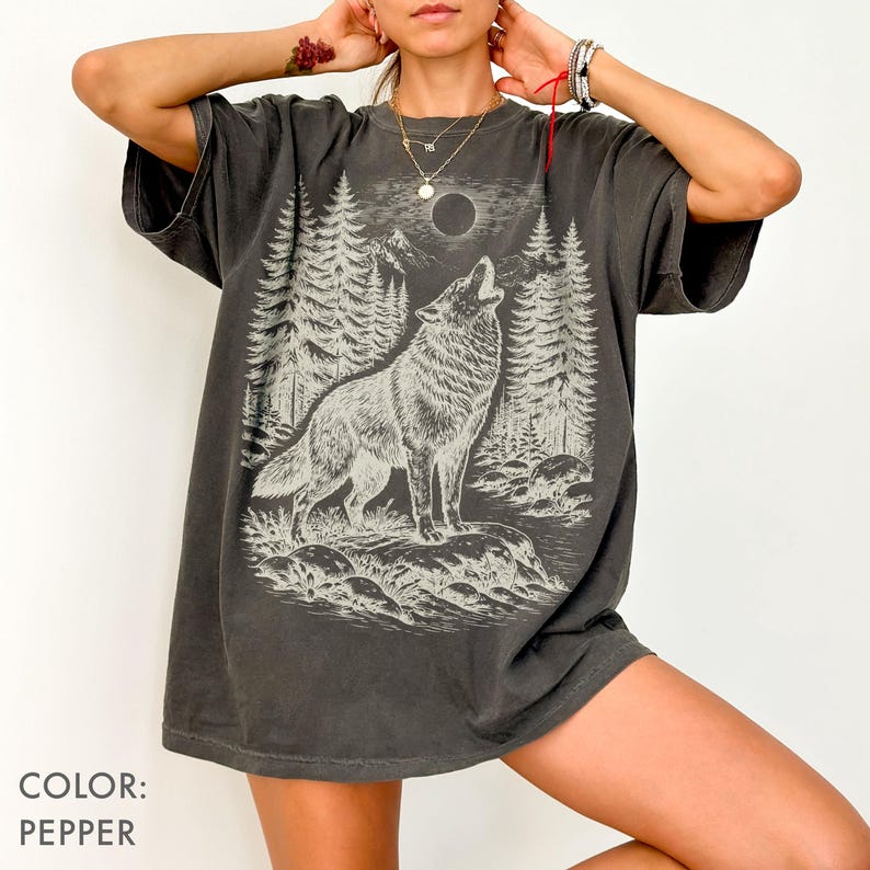 Puede incluir: Una camiseta color pimienta con un estampado gr&aacute;fico de un lobo aullando a la luna llena en un entorno forestal. El dise&ntilde;o incluye ilustraciones detalladas de pinos, terreno rocoso y una luna circular, representadas en tonos claros y apagados.