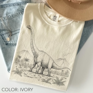 Pode incluir: Camiseta de cor marfim com uma ilustração detalhada de um dinossauro Brachiosaurus em uma paisagem pré-histórica. O design inclui montanhas, palmeiras e samambaias. A camiseta está dobrada, com um chapéu e jeans visíveis.