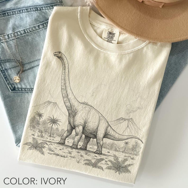 Ivory Comfort Colors unisex oversized t-shirt featuring a vintage-style Brontosaurus dinosaur illustration with volcanoes, palm trees, ferns, and prehistoric landscape details in a pencil sketch graphic style.
