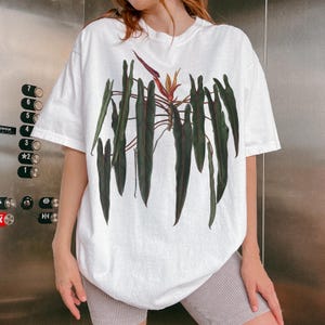 May include: A white short-sleeve t-shirt featuring a large botanical illustration of a philodendron plant with long, dark green, pointed leaves and a single orange and purple flower centered on the chest area of the garment.