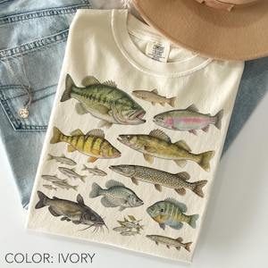 Könnte beinhalten: Elfenbeinfarbenes T-Shirt mit einer detaillierten Illustration verschiedener Fischarten. Das Design umfasst einen Schwarzbarsch, eine Forelle, einen Barsch und andere Fische. Das Shirt wird mit blauen Jeans und einem beigen Hut kombiniert.