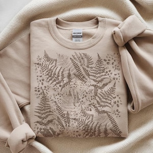 Puede incluir: Sudadera beige de cuello redondo con un dise&ntilde;o de helechos en tonos marrones. La sudadera est&aacute; hecha de un material suave y tiene una etiqueta que dice "GILDAN HEAVY COTTON". El dise&ntilde;o de helechos est&aacute; impreso en la parte delantera.