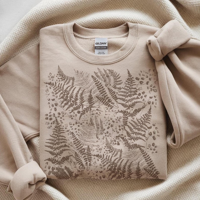 Puede incluir: Sudadera beige de cuello redondo con un dise&ntilde;o de helechos en tonos marrones. La sudadera est&aacute; hecha de un material suave y tiene una etiqueta que dice "GILDAN HEAVY COTTON". El dise&ntilde;o de helechos est&aacute; impreso en la parte delantera.