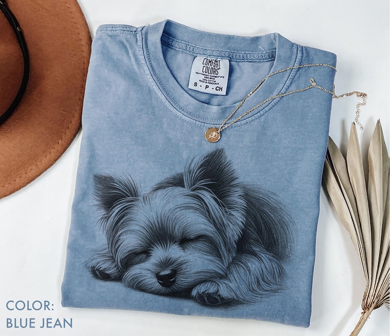 Puede incluir: Camiseta azul vaquero con una ilustraci&oacute;n detallada de un Yorkshire Terrier durmiendo. La camiseta tiene cuello redondo y mangas cortas. Un collar de oro con un colgante descansa sobre la camiseta. La etiqueta dice "Comfort Colors".