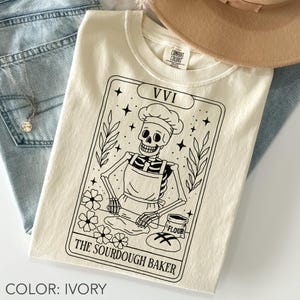 May include: Ivory-colored t-shirt featuring a black and white graphic of a skeleton baker in a chef's hat and apron, rolling dough. The design is framed with stars, flowers, and the text "The Sourdough Baker" and "VVI".
