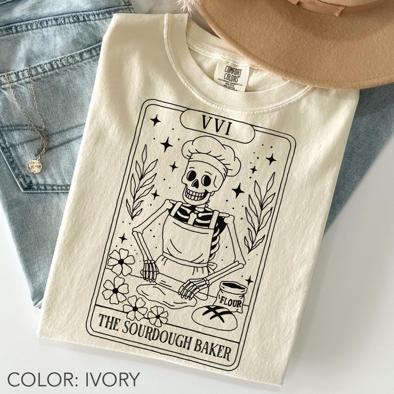 May include: Ivory-colored t-shirt featuring a black and white graphic of a skeleton baker in a chef's hat and apron, rolling dough. The design is framed with stars, flowers, and the text "The Sourdough Baker" and "VVI".