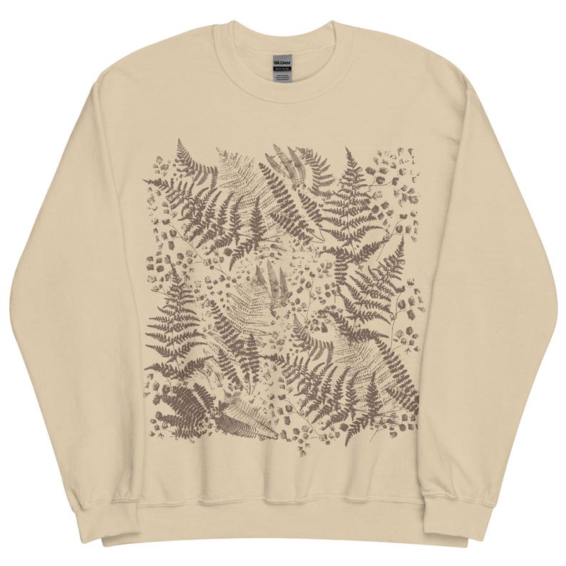 Puede incluir: Sudadera beige con un estampado de helechos marrones. El dise&ntilde;o es un patr&oacute;n repetido de ilustraciones detalladas de helechos, creando un tema natural y bot&aacute;nico. La sudadera tiene cuello redondo y mangas largas, adecuada para uso casual.